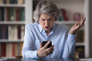 Angry older businesswoman sit at workplace staring at cellphone screen, feels mad looks shocked by...