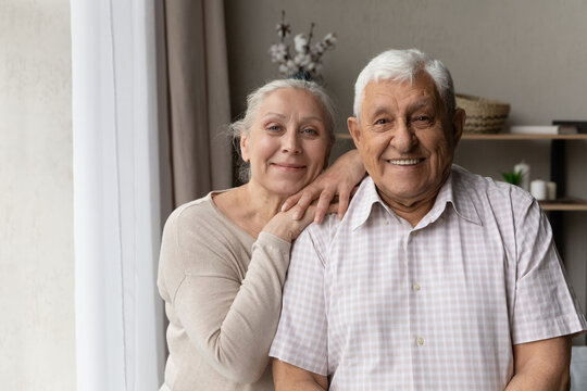 Portrait of grey-haired grandparents smile staring at camera feel satisfied, standing in living room, spend carefree retired life together. Happy marriage, pure love, good harmonic relations concept