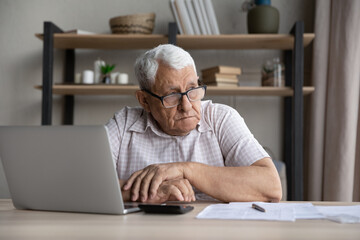 Pensive anxious older man in glasses sit at table with heap of bills, calculator and laptop, manage...