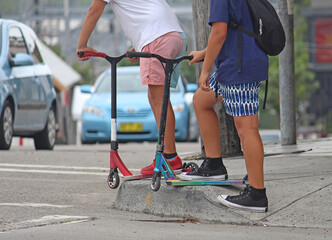 Obraz premium Two teenage boys at an intersection on scooters waiting for the traffic lights to change. Cars in the background. Generation Z.