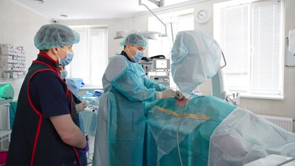Middle-aged plump doctor inserts sharp needle-like instrument into patient's body. Assistants stand behind waiting for instructions.