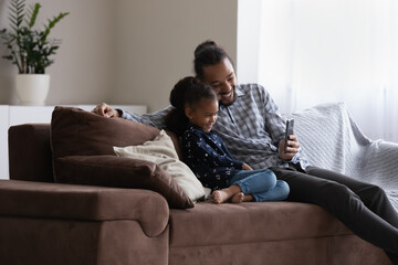 Cheerful little kid girl enjoying leisure time with daddy. Happy father taking selfie photo on mobile phone with beloved adorable daughter, resting on couch, talking on video call on smartphone