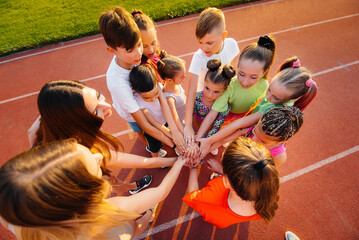 A large group of children, boys and girls, stand together in a circle and fold their hands, tuning up and raising team spirit before the game at the stadium during sunset. A healthy lifestyle.