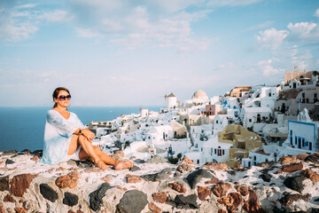 Fototapeta na wymiar Junge Frau mit Sonnenbrille sitzend auf einer Mauer und geniesst die Sonne auf der Insel Santorin in Griechenland