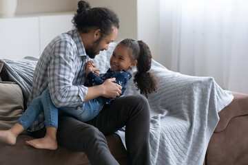 Playful happy African dad tickling cheerful little daughter kid, cuddling child on couch with love,...