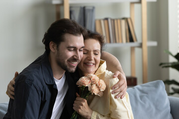 Grateful happy mature mom embracing grown son man with gratitude, love, holding flowers bouquet,...