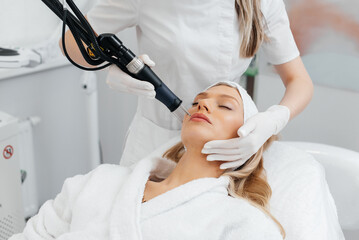 Naklejka premium A young girl gets a charcoal peeling for the skin of the face in a beauty salon. Laser pulses cleanse the skin. Hardware cosmetology. The process of photothermolysis, warming the skin.