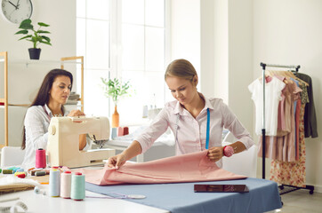 Obraz premium Women tailors work in own modern fashion atelier sew garment on sewing machine. Female seamstresses or dressmakers design new clothing line in workshop. Style concept. Dressmaking and manufacturing.