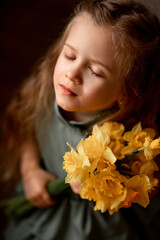 Naklejka premium beautiful spring portrait of a cute little girl with long hair and big green eyes. and a bouquet of yellow daffodils in her hand. The child enjoys the flowers and closed her eyes