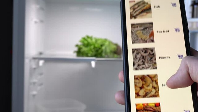 A man stands in front of an open empty refrigerator and scrolls through an assortment of online store products.