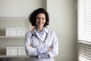 Confident happy mature chief doctor woman in white coat standing with hands folded in office,...