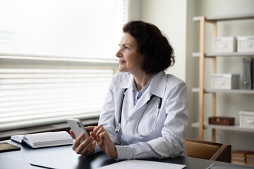 Happy thoughtful mature doctor woman using online app on mobile phone, consulting internet, texting message on chat, making appointment with patients, looking away at window, thinking at work table