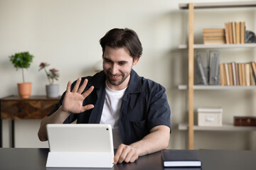 Happy friendly freelance tablet user guy waving hand hello at gadget screen, making distant video call to client, customer, using portable pc for business communication at office workplace table