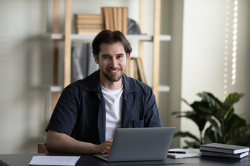 Fototapeta premium Happy successful young business professional man in casual typing on laptop, working in home office, looking at camera, smiling. Millennial businessman, freelance employee head shot portrait