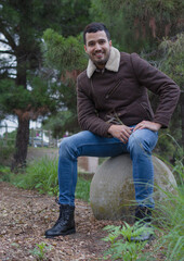 Naklejka na ściany i meble full length shot of a latin guy smiling and looking at the camera while is sitting on a concrete sphere in the woods
