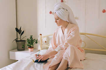 Young happy business woman with a towel on her head lies in bed in pajamas in a cozy home with notebook. Social media. Beauty routine. Skin care cosmetic concept