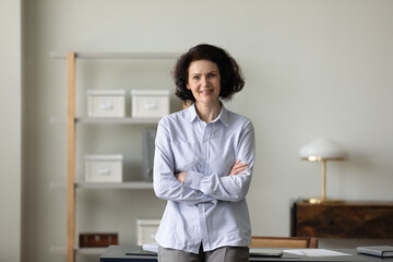 Confident mature business leader woman standing with hands folded at office workplace, looking at camera, smiling. Senior businesswoman, CEO, entrepreneur head shot portrait