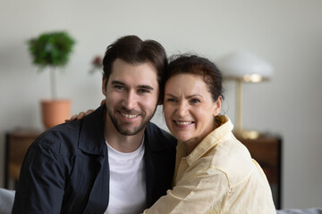 Obraz na płótnie Canvas Happy mature mom embracing grown handsome son at home, looking away with toothy smile, posing for camera. Millennial guy enjoying family leisure time, meeting with mature mother. Head shot portrait