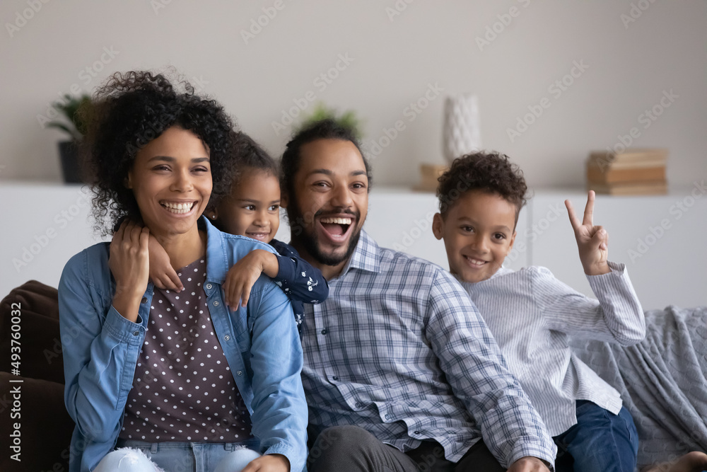 Wall mural Excited joyful Black family with two cute sibling boy and girl having fun together at home, resting on couch, laughing, shouting, showing victory. African parents and little kids portrait