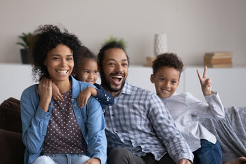 Excited joyful Black family with two cute sibling boy and girl having fun together at home, resting on couch, laughing, shouting, showing victory. African parents and little kids portrait