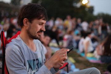 A man sits in a camping chair at an open-air music festival and looks at his phone