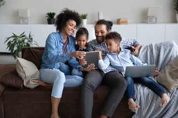 Cheerful young Black parents and curious little gen Z boy and girl resting together on home sofa, using laptop, tablet computer gadget, shopping internet, chatting online on social media