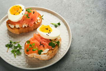 Sandwich with delicious toppings smoked salmon, eggs, herbs and microgreens radish, black sesame seeds over white plate on gray concrete table background. Healthy open sandwich superfood. Top view.