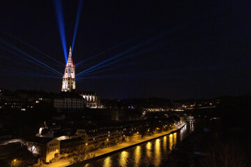 The cathedral in Bern, Switzerland, illuminated during Museumsnacht 2022, the night of museums. Several floodlights on the walls of the bell tower create rays of light in the dark sky. The Aare River