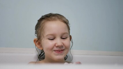 A lovely European preschooler girl takes a bath cheerfully blowing foam from her palms and smiles beautifully. Slow motion. Close-up