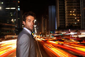 A city boy through and through. A portrait of a handsome businessman standing in the middle of a busy city street at night.