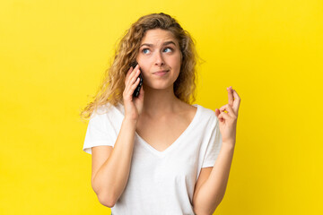Young blonde woman using mobile phone isolated on yellow background with fingers crossing and wishing the best