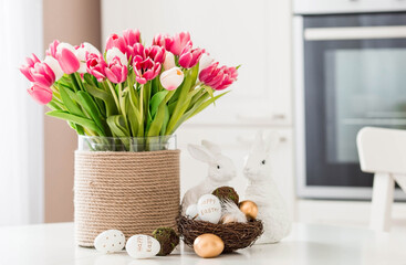 A bouquet of tulips, Easter bunnies and eggs with a golden pattern on the table. In the background is a white Scandinavian-style kitchen. Beautiful greeting card. The minimal concept.