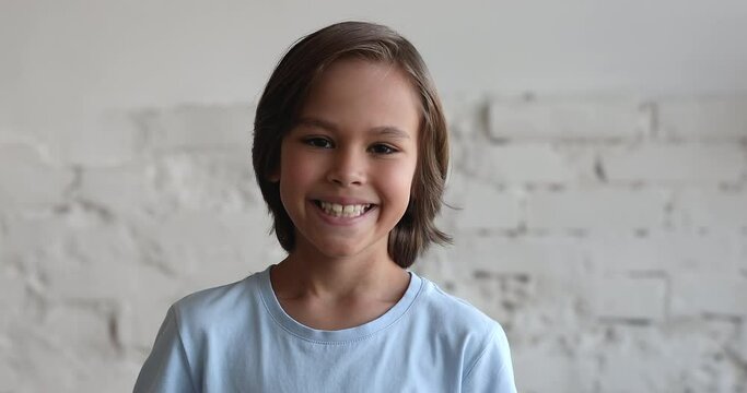 Head shot of handsome tanned pre-teen 10s boy standing alone against white brick wall background, staring at camera, looks happy. Portrait of joyful candid child with wide toothy smile, gen Z concept