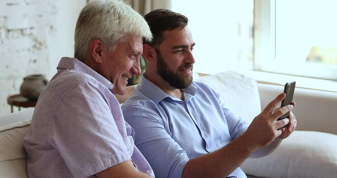 Close up smiling older man and adult 35s son sit on sofa with smartphone, have fun, enjoy new mobile application looking happy spend time together on weekend at home. Leisure using modern tech concept