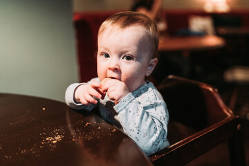 Baby girl sitting at the table on a high chair in a pub, eating. Baby portrait.