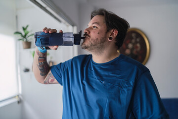 hombre con camiseta azul y tatuajes en el brazo bebiendo agua. hombre de estilo alternativo bebiendo de una botella de plástico color azul