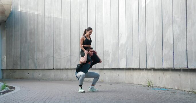 Strong athletic man doing squatting with woman sitting on his shoulders. Sports couple train together. Fitness workout training outdoors