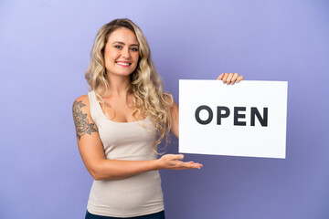 Young Brazilian woman isolated on purple background holding a placard with text OPEN with happy expression