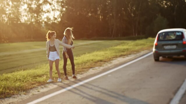 Pretty women travelers waiting for car s at roadside. Young sexy girls dressed in casual summer clothes hitchhiking on the road raising their thumbs up. Concept of freedom and travelling.