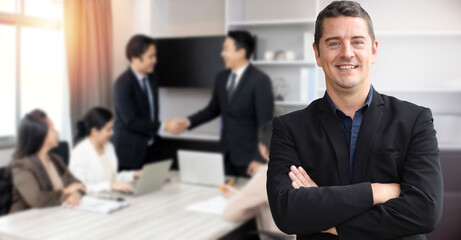 Fototapeta na wymiar Smart and smiling handsome caucasian businessman standing and arms cross with confident on background of business people shaking hands after contract signed in modern office. Success business concept