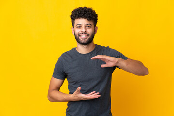 Young Moroccan man isolated on yellow background holding copyspace imaginary on the palm to insert...