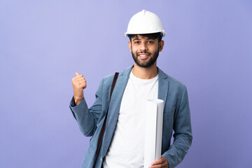 Young architect Moroccan man with helmet and holding blueprints over isolated background pointing back
