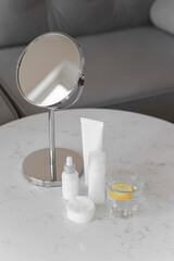 White bottles of skincare cosmetics on a marble table. A glass of water with lemon and mirror.