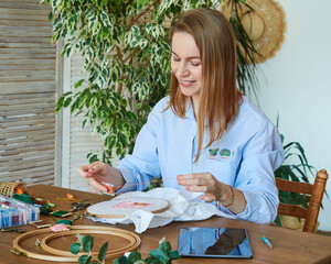 Happy blonde woman cross-stitch, sitting at a wooden table, uses a computer tablet