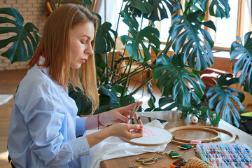 Cute blonde girl in a blue shirt is cross-stitching while sitting at a wooden table