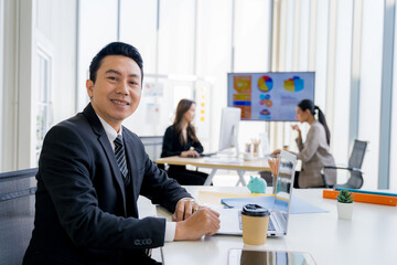 Attractive asian young confident businessman sitting at the office table with group of colleagues in the background, working on laptop computer