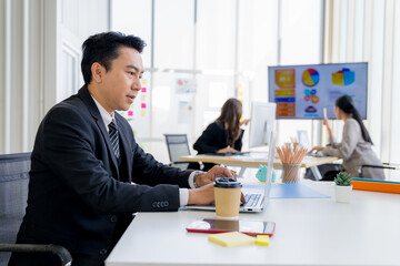 Attractive asian young confident businessman sitting at the office table with group of colleagues in the background, working on laptop computer