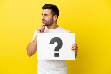 Young handsome caucasian man isolated on yellow background holding a placard with question mark...