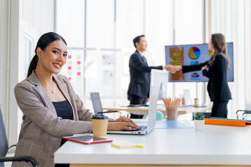 Attractive confident confident Asian businesswoman sitting at office desk with group of colleagues holding hands in the background. Works on laptop and tablet computers.