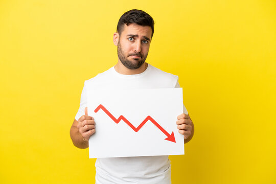 Young handsome caucasian man isolated on yellow background holding a sign with a decreasing statistics arrow symbol with sad expression